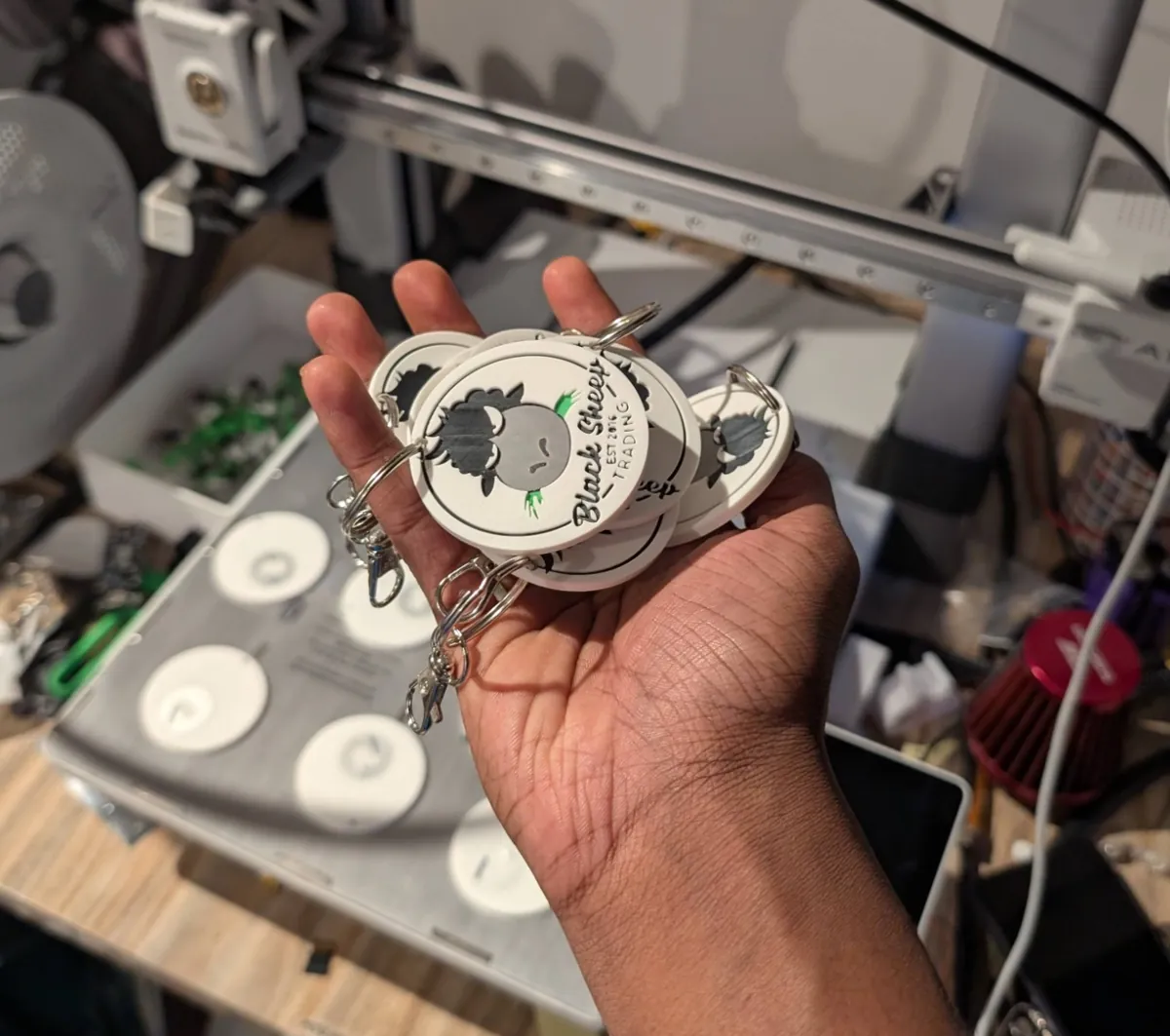 A handful of printed Black Sheep Trading keychains in front of the printer, with more still on the bed