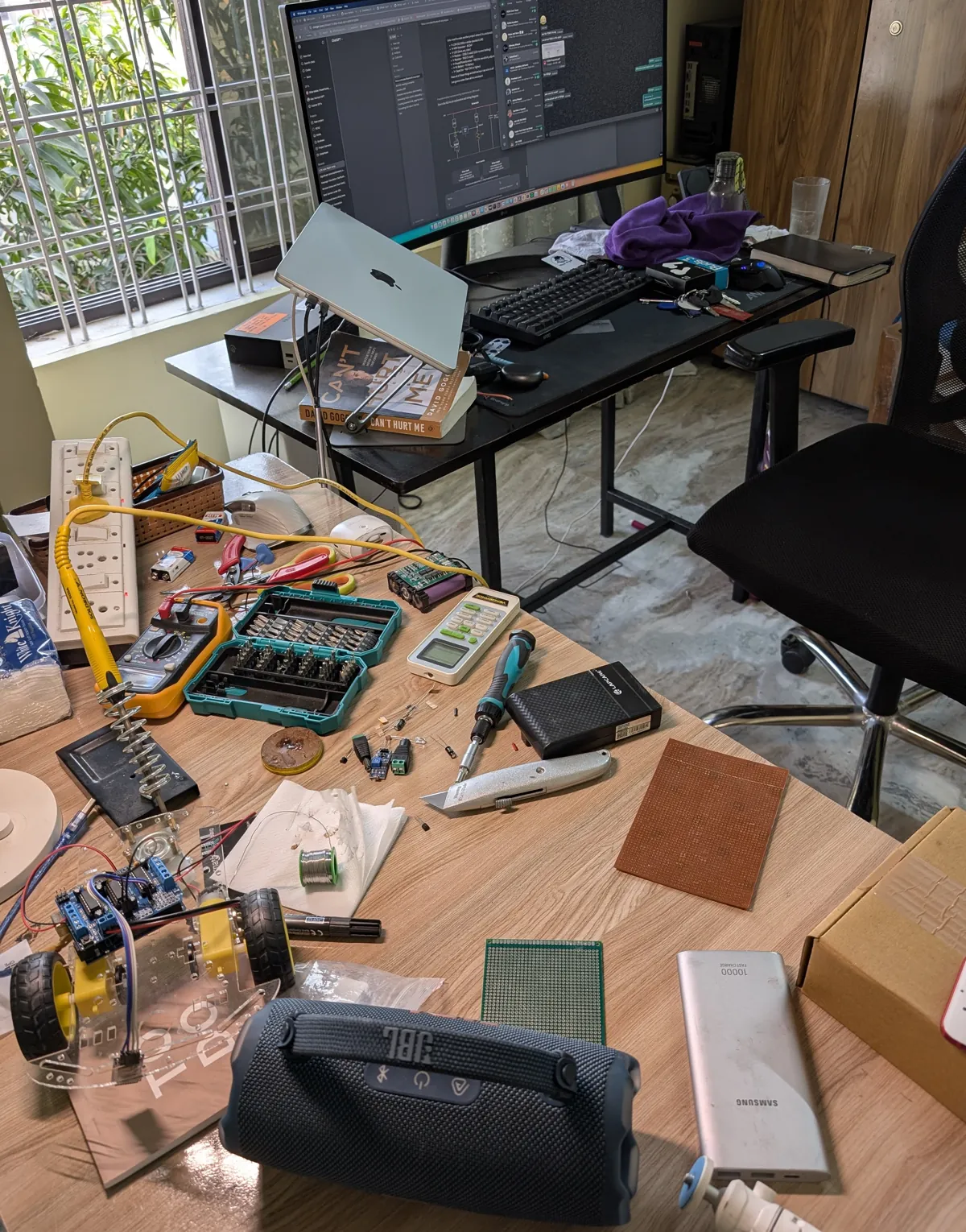 A work desk in Chennai: multimeter, screwdriver set, a small robot chassis, perfboard, solder and a monitor with a workflow editor open