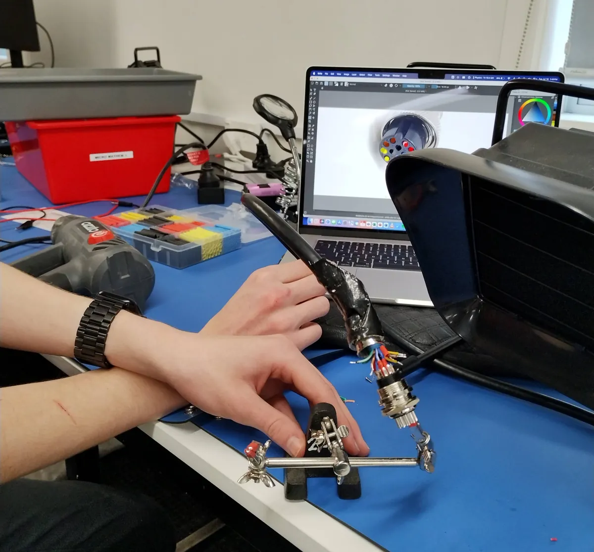 Soldering a multi-pin harness connector in a helping-hands clamp, laptop with the CAD model behind